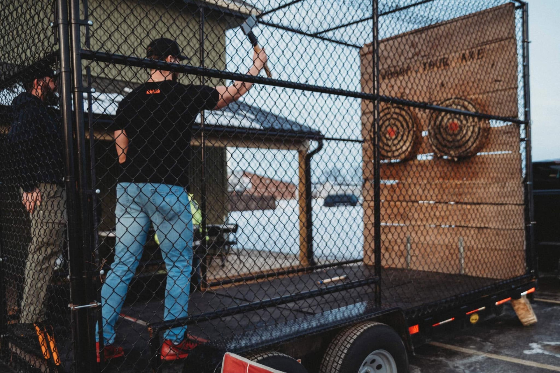 Men throwing axes in a caged trailer range at dusk